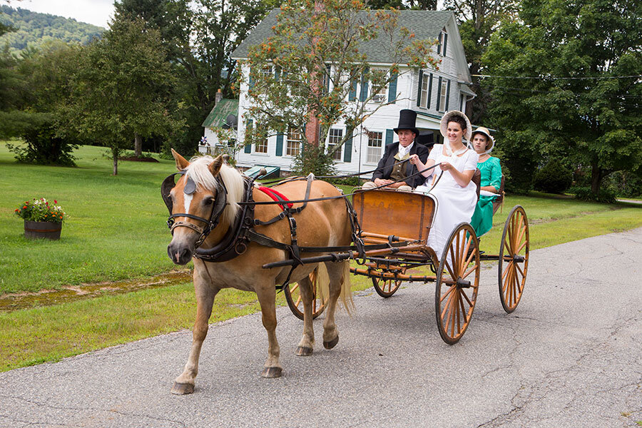 Women in white gown and bonnet driving a horse-drawn carriage through the village