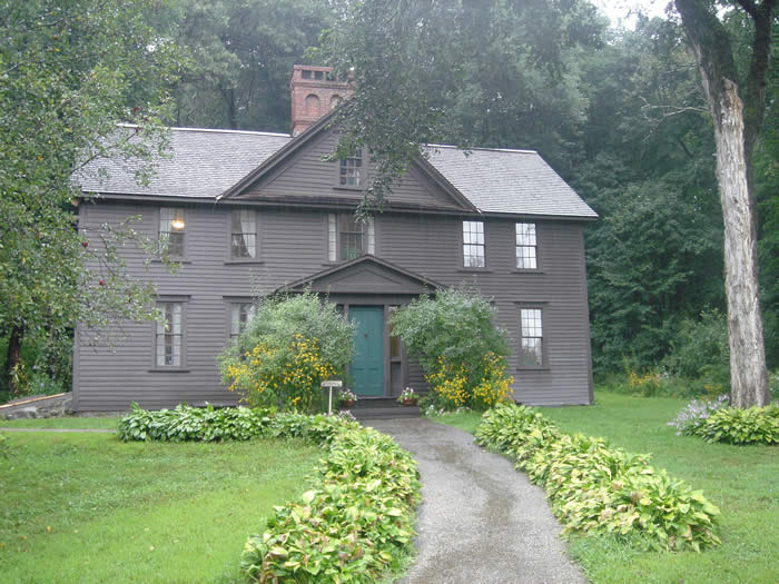 Brown clapboard 19th Century house flanked by tall grees with a wide path through the lawn to the green front door