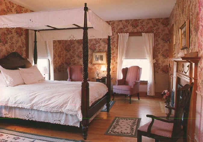 Queen-sized canapy four-poster bed in room with pink floral wallpaper and two wing chiars 